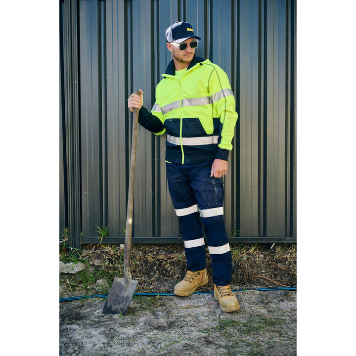 Person wearing high-visibility workwear, wearing a Bisley BCAP50 Cap, holding a shovel against a metal wall.