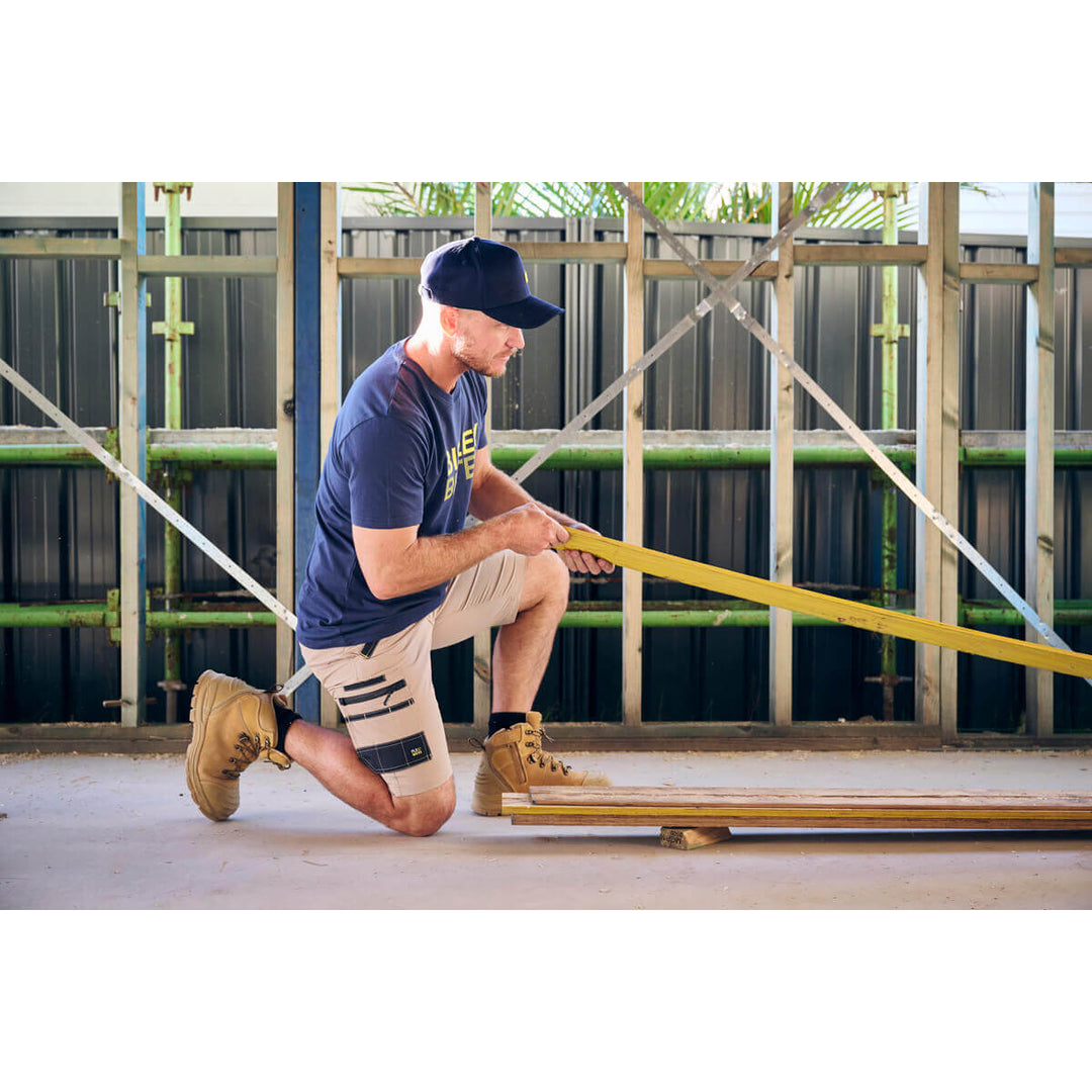 Man kneeling on a construction site using a long tool, wearing a Bisley BCAP50 Cap.