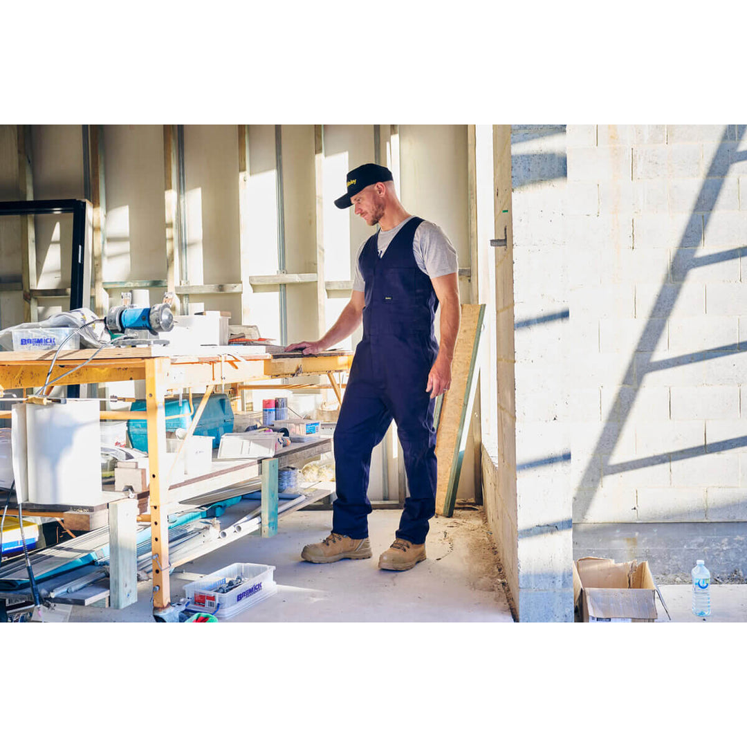 Man in blue action back overalls, wearing a Bisley BCAP50 Cap, working on a construction site with tools and materials around.