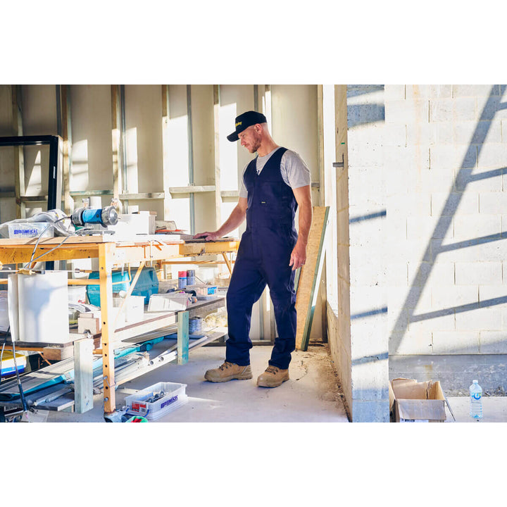 Man in blue action back overalls, wearing a Bisley BCAP50 Cap, working on a construction site with tools and materials around.